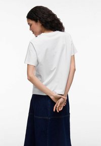 Witte katoenen t-shirt met een ontspannen pasvorm, korte mouwen en een rechte zoom. Het model draagt een donkere spijkerrok, met de handen samengevouwen achter. 