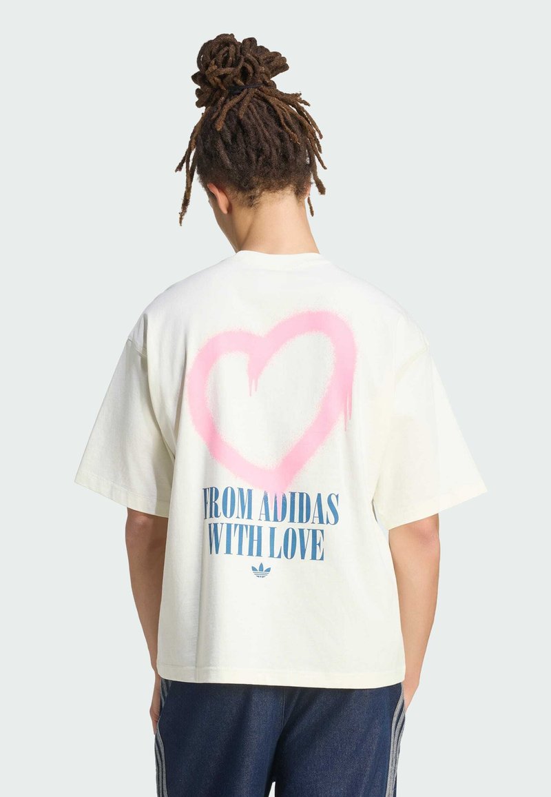 Person mit Dreadlocks trägt ein weißes T-Shirt mit großem pinkfarbenem Herz und der Aufschrift "FROM ADIDAS WITH LOVE" auf der Rückseite, kombiniert mit dunkler Hose.