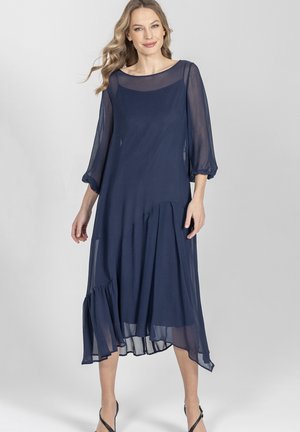 Kobieta ubrana w granatową, przezroczystą sukienkę midi z długim rękawem, asymetrycznym falbanistym dołem oraz czarne szpilki na pasku, stojąca na jednolitym tle.