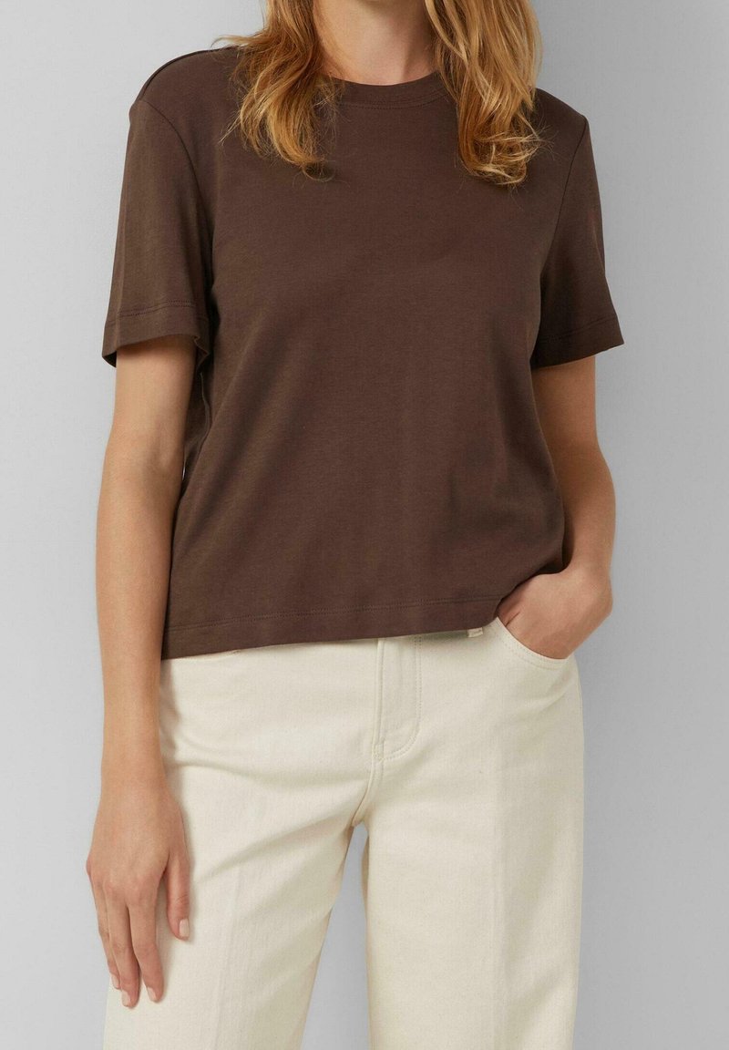 Personne portant un t-shirt marron uni à manches courtes et un pantalon beige clair, debout avec la main droite dans la poche et le bras gauche détendu.