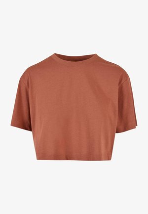 Osoba w rdzawym krótkim T-shircie i czarnych, wysokich dżinsach z rękami w kieszeniach na jednolitym tle.