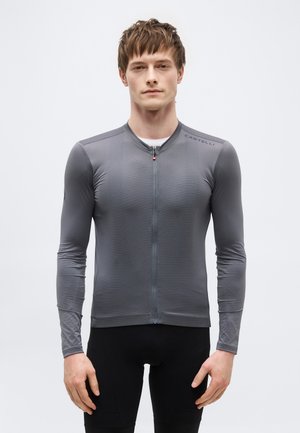 Ung mann iført en tettsittende, langermet grå sykkeltrøye med frontglidelås og "Castelli"-logo på skulderen, stående foran en ensfarget bakgrunn.