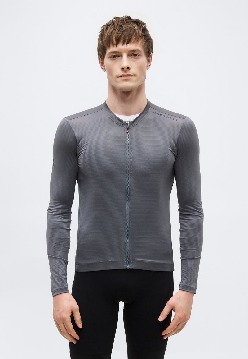 Νεαρός άνδρας που φοράει εφαρμοστό μακρυμάνικο γκρι ποδηλατικό τζέρσεϊ με φερμουάρ μπροστά και λογότυπο "Castelli" στον ώμο, στέκεται μπροστά σε απλό υπόβαθρο.