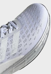 Zapatilla de running gris claro con un upper de punto texturizado, acentos de rayas blancas y una suela de goma con un patrón de banda de rodadura prominente.