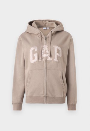 Beige zip-up hoodie made of soft fabric, featuring a large "GAP" logo in a lighter beige, with a front pocket and adjustable hood.
