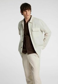 Giacca button-up beige chiaro con due tasche frontali, abbinata a una camicia marrone scuro e pantaloni beige chiaro. Il tessuto ha un aspetto testurizzato e intrecciato.