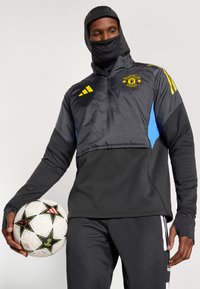 Schwarze und graue Sportjacke mit blauen Akzenten, mit Reißverschluss am Kragen und Kapuze. Hat gelbe Streifen und das Manchester United-Logo. Schwarze Hose und ein Fußball.
