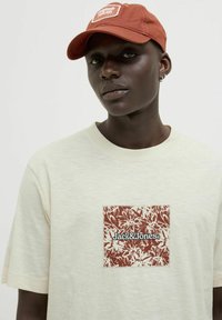 Šviesiai bežinė trumpomis rankovėmis marškinėliai su tekstūruotu rudos spalvos gėlių aplikacija ir juodu "Jack & Jones" logotipu; derinami su rūdžių spalvos kepure.