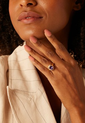 Gouden ring met een centrale paarse edelsteen omringd door kleinere kristallen, gedragen op een hand met nude matte nagels, tegen een lichte stofachtergrond.