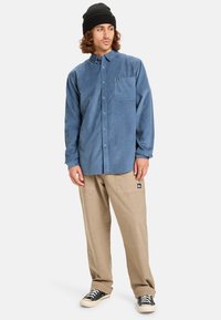 Blauwe corduroy knoopshirt met een borstzak, gecombineerd met beige cargobroeken met zijzakken en een logo-opdruk. Zwarte beanie en sneakers.