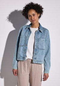 Chaqueta de denim azul claro con cuello clásico, dos bolsillos frontales y cierres a presión. Combinada con una camisa blanca y pantalones beige.