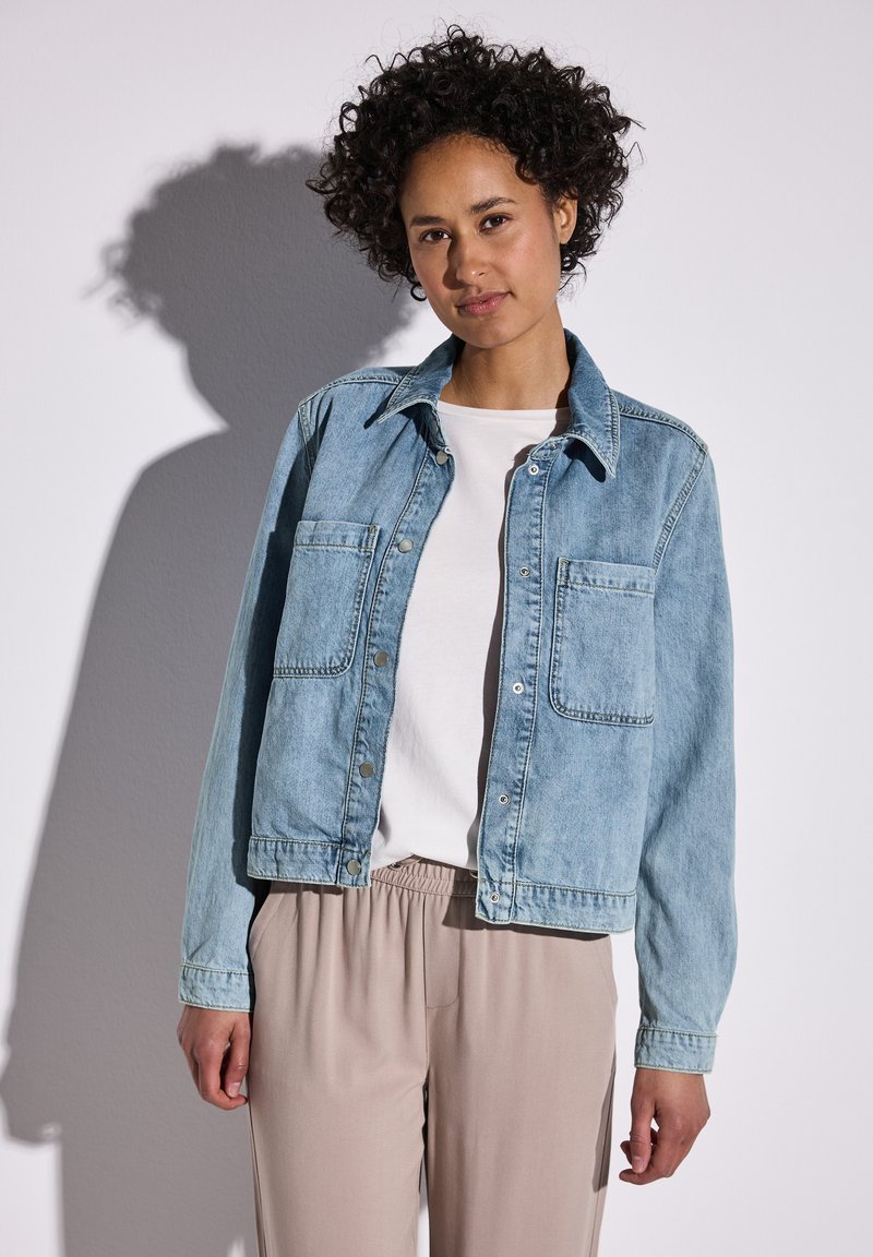 Chaqueta de denim azul claro con cuello clásico, dos bolsillos frontales y cierres a presión. Combinada con una camisa blanca y pantalones beige.