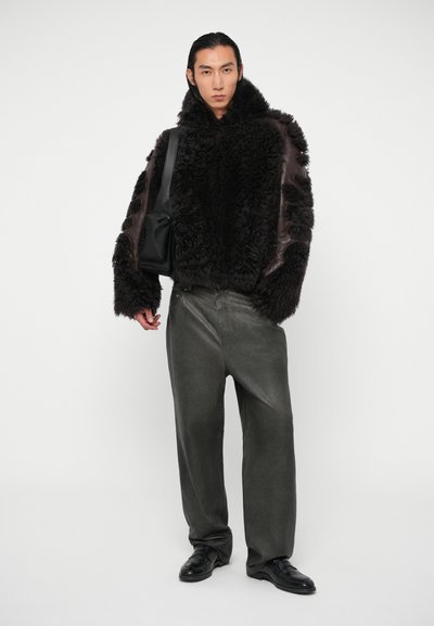 Chaqueta forrada de piel negra con acentos de cuero marrón, pantalones grises y zapatos negros. El modelo está de pie contra un fondo blanco, sosteniendo un bolso.