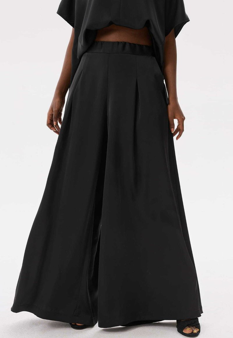 Vrouw in hoge taille, wijde zwarte broek met zijzakken en een zwart crop top, staand tegen een effen achtergrond.