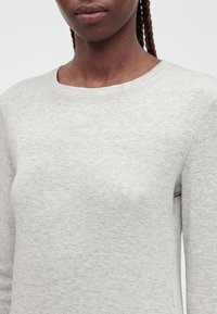 Joven con piel oscura y cabello trenzado que lleva una camiseta gris claro de manga larga y cuello redondo, mostrado desde los hombros hasta la boca.