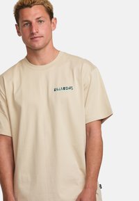 Beige short-sleeve T-shirt made of cotton, featuring a printed logo in dark green on the left chest area, with a relaxed fit and standard crew neckline.