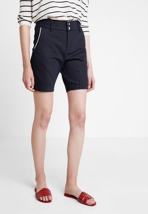 Kvinde iført sorte shorts med hvide sidelister, en langærmet top med striber og røde åbne sandaler, der står på et hvidt gulv.