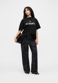 Γυναίκα με σγουρά μαλλιά που φοράει μαύρο t-shirt AllSaints, σκούρο παντελόνι φαρδύ με λεοπάρ σχέδιο, κρατώντας μαύρο τσαντάκι, στέκεται σε λευκό φόντο.