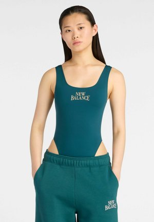 Ung kvinde iført teal New Balance badedragt og matchende træningsbukser, stående med hænderne i lommerne mod en ensfarvet baggrund.