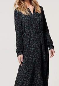 Femme portant une robe noire à manches longues avec un petit motif floral vert, boutonnée devant, ceinturée à la taille.