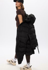 Cappotto puffo nero con grandi sezioni trapuntate, chiusura con zip e lacci. Indossato sopra una maglia marrone e abbinato a sneakers beige.