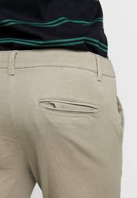 Pantaloni beige con una texture liscia, dotati di una piccola tasca posteriore orizzontale e passanti per la cintura. La vita è semplice, senza attrezzature visibili.