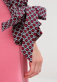 Mönstrad tygrosett med röd blomdesign på vit bakgrund, fäst vid ett rosa plagg, med flödande band.