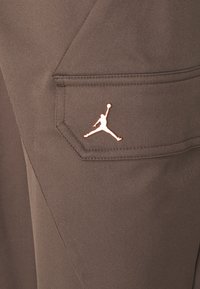 Ruskeat urheiluhousut, joissa on sivutasku ja metallinhohtoinen ruusukultainen Jumpman-logo. Sileä, kestävä kangas, jossa on tekstuuri.