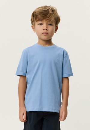 Ung gutt med lysbrunt hår iført en lys blå t-skjorte med oppkvelte ermer og mørke shorts, stående mot en ensfarget lys bakgrunn.