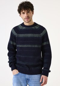 Suéter de punto azul marino con rayas verdes horizontales, textura acanalada, escote redondo y mangas largas. Combinado con jeans de color azul claro.