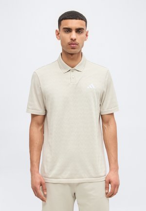 Νέος άνδρας με κοντά σκούρα μαλλιά, που φοράει μπεζ πόλο πουκάμισο Adidas και αντίστοιχο παντελόνι, στέκεται μπροστά σε ανοιχτόχρωμο απλό φόντο.