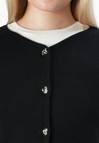 Gros plan sur un cardigan noir avec des boutons abstraits en argent brillant porté sur un haut côtelé blanc, sur une personne aux longs cheveux blonds.