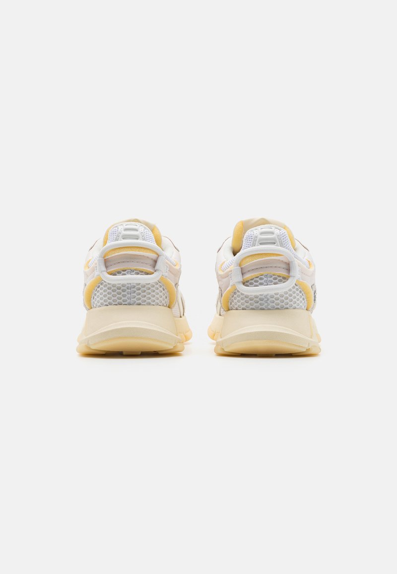 Zapatillas con una parte superior de malla blanca, acentos amarillos y una suela gruesa color crema. Presenta un collar acolchado y una tira de tira para facilitar su uso.
