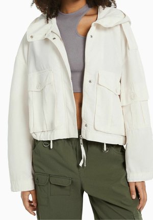 Kvinde iført en kort hvid lynlåsjakke med klaplommer, grå ribbet crop top og olivengrønne cargobukser med flere lommer.