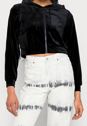 Zbliżenie osoby noszącej czarną aksamitną krótką bluzę z kapturem na suwak i białe spodnie z wysokim stanem z czarnymi paskami tie-dye.