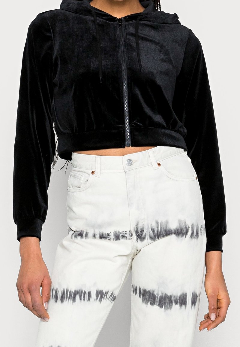 Gros plan d'une personne portant un sweat à capuche court en velours noir avec fermeture éclair et un pantalon blanc taille haute avec des rayures tie-dye noires.