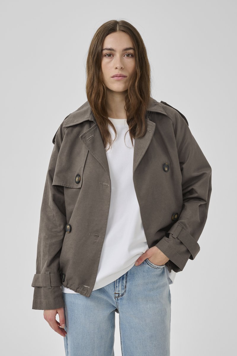 Frau mit langen braunen Haaren, die eine graue zweireihige Jacke, ein weißes T-Shirt und hellblaue Jeans trägt, steht vor einem einfarbigen Hintergrund.