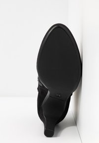 Bottine en daim noir à bout pointu avec un talon haut et fin, vue de la semelle sur un fond blanc en angle.