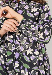 Camicia nera a fiori con un motivo di fiori bianchi, viola e verdi. Design con bottoni, maniche lunghe e tessuto текстurizzato.