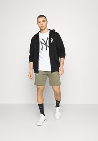 Sudadera negra, camiseta blanca con logo de NY, pantalones cortos verdes oliva, calcetines negros y zapatillas blancas. Fondo liso, enfoque en ropa deportiva casual.