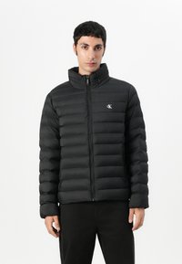 Must ümmarguste õmblustega jope kõrge krae ja esikäe tõmblukuga. Omab horisontaalset tepitud mustrit ja väikest logo rinnal.