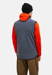 Fleece-Weste in Grau mit schwarzem Besatz, mit orangefarbenem Kapuzenoberteil und kontrastierenden Ärmeln. Getragen mit hellbraunen Hosen, minimalen Hardware-Details.