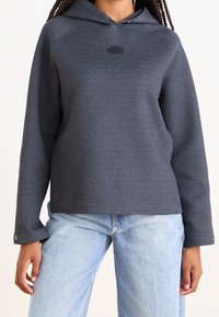 Donkergrijze hoodie met een logo aan de voorkant, raglanmouwen en een rechte zoom. Gecombineerd met lichtblauwe jeans met knoopmanchetten.