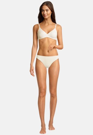 Donna con capelli castani lunghi che indossa un reggiseno color crema e biancheria intima coordinata, in piedi davanti a uno sfondo neutro.