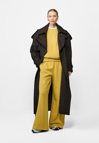 Czarny płaszcz trenczowy na żółtej bluzie i pasujących szerokich spodniach. Gładki materiał o luźnym kroju i kontrastujących odcieniach. Sportowe buty.