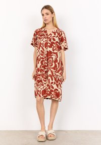 Short-sleeve beige dress featuring a large red floral pattern, a v-neckline, and a loose, knee-length fit. Worn with neutral sandals.