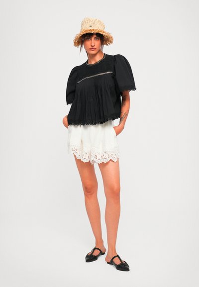Woman wearing a black puff-sleeve top with lace trim, white lace-hem shorts, black flat shoes, and a tan woven straw hat, standing with hands in pockets.