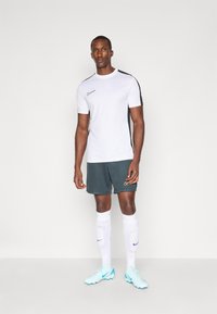 Nike sportska odjeća koja se sastoji od bijele majice kratkih rukava s crnim detaljima, u kombinaciji s tamnozelenim šortsevima i svijetloplavim kopačkama.