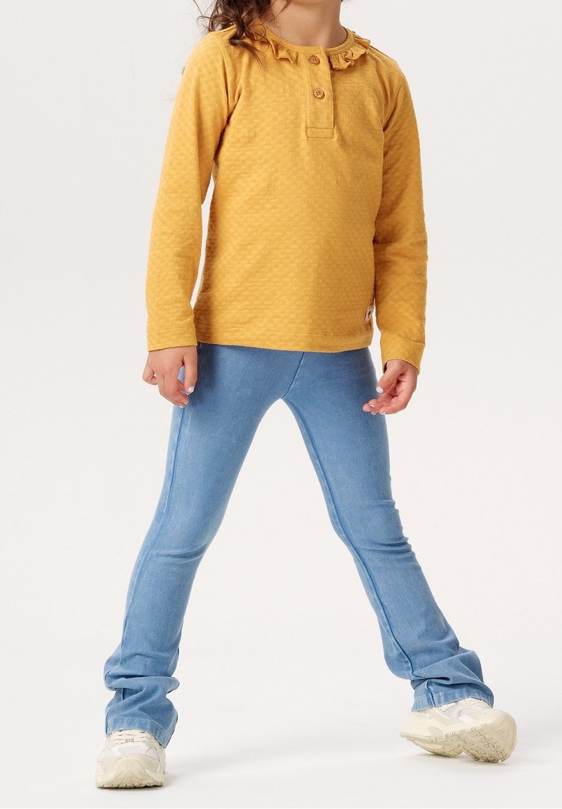 Enfant portant un haut jaune à manches longues avec des volants et un jean bleu clair, debout, jambes écartées, portant des baskets blanches.