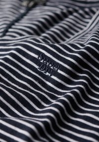 Marineblå og hvidstribet stof med broderet tekst "Super Dry" i mørk tråd, der viser folder og tekstur.